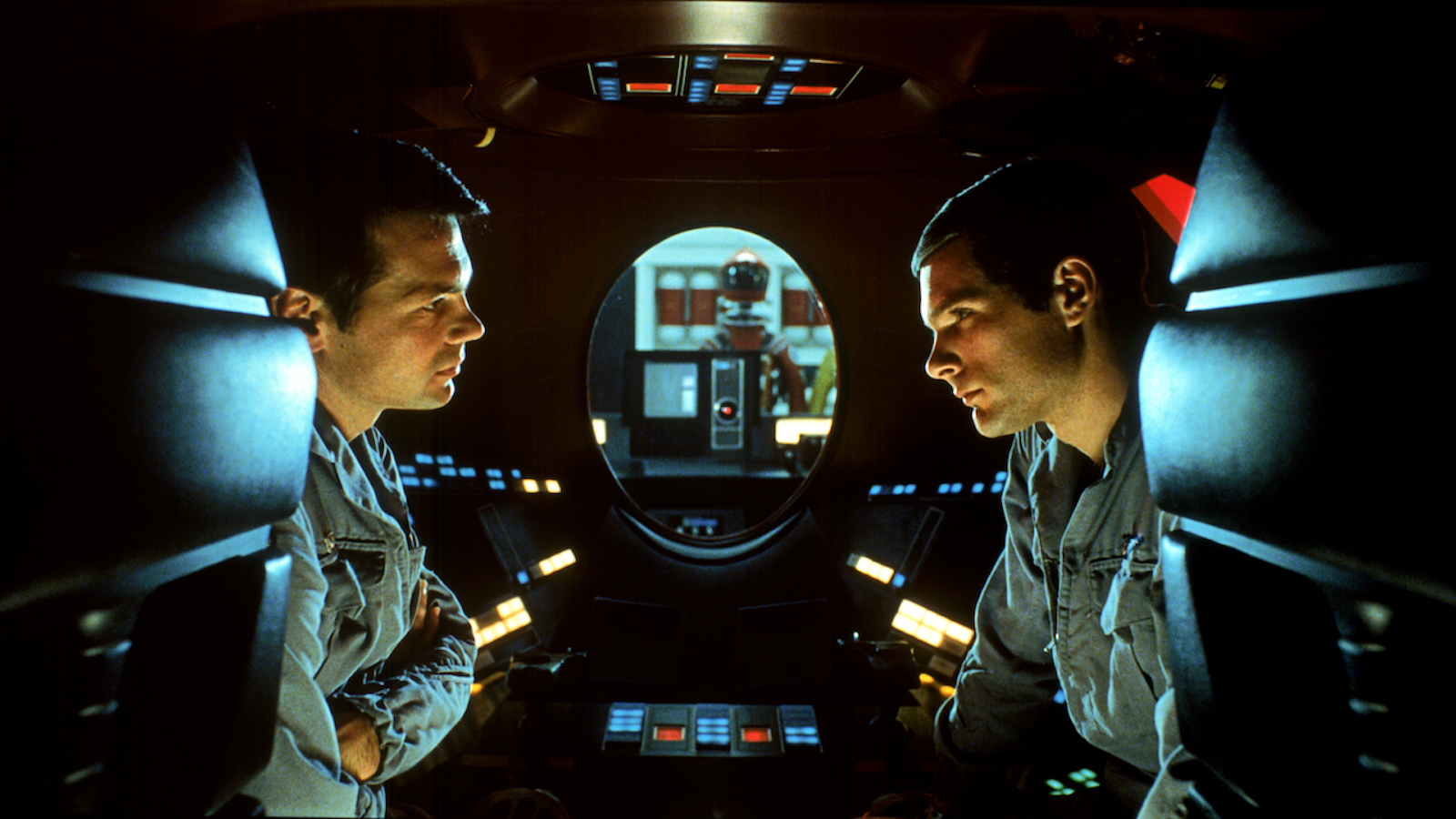 Two astronauts in profile sit in an enclosed pod and discuss with an oval shaped window behind them showing a computer's red light pointed in the direction of the camera