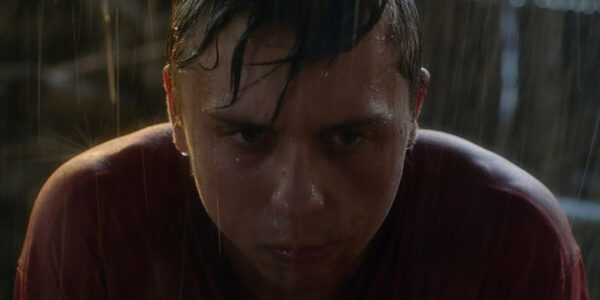 A boy in close-up with an intense face staring straight ahead covered in pouring rain