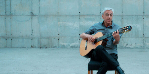 A white haired man holds a guitar as he sits in a chair set against a large grey cement wall