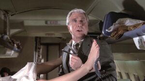 In an airplane aisle, a goofy white-aired man grips forceps while looking up from two legs in stirrups.