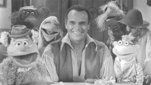 A black and white image of a man smiling surrounded by Muppets, all looking at camera