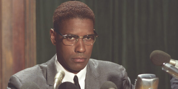 A man wearing spectacles and a serious expression sits in front of a bunch of press microphones