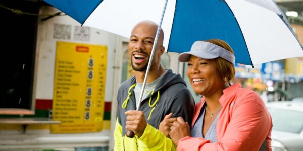 A man in yellow and gray windbreaker and a woman in a pink jacket stand under a blue and white umbrella in the rain smiling