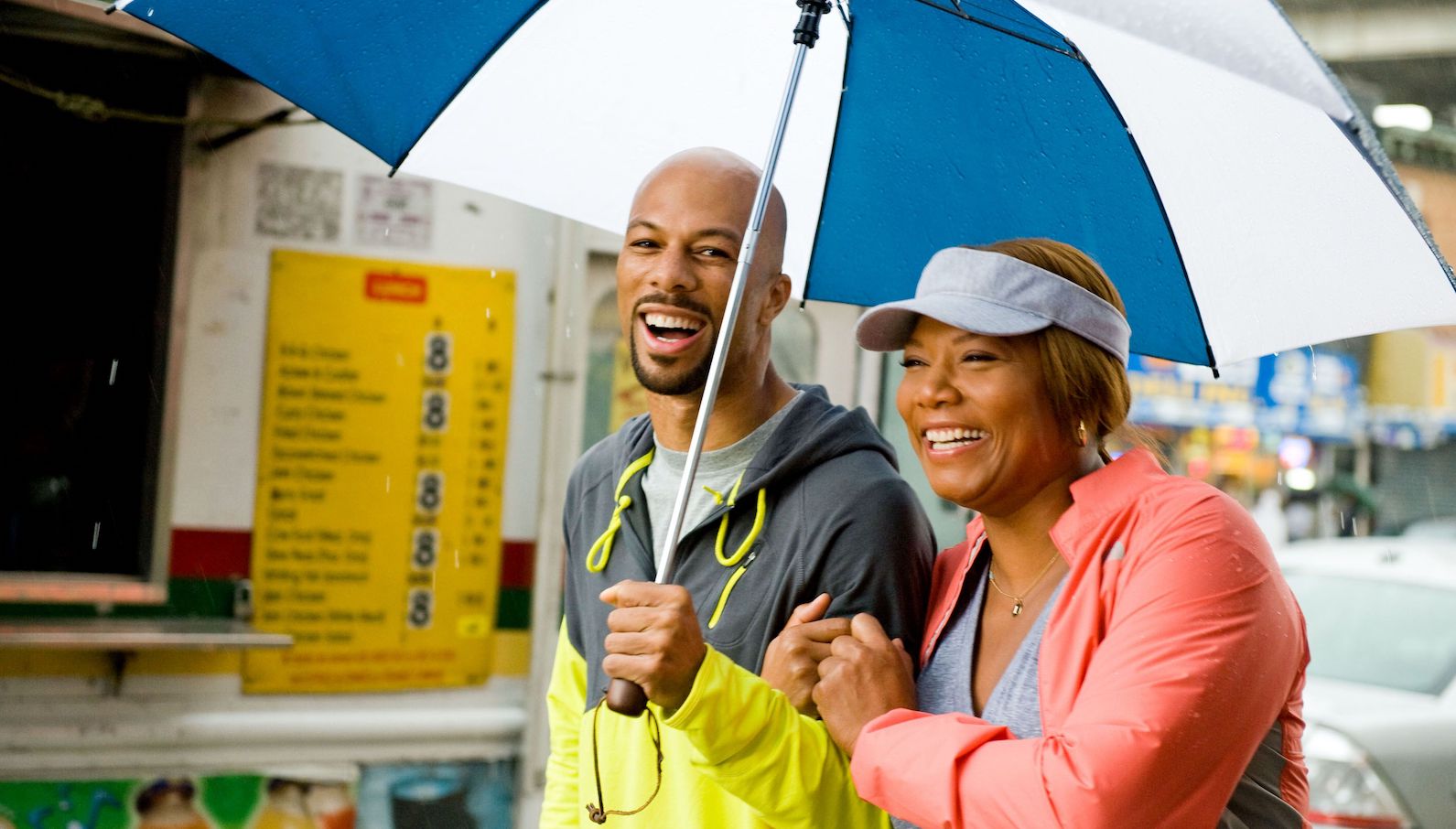 A man in yellow and gray windbreaker and a woman in a pink jacket stand under a blue and white umbrella in the rain smiling