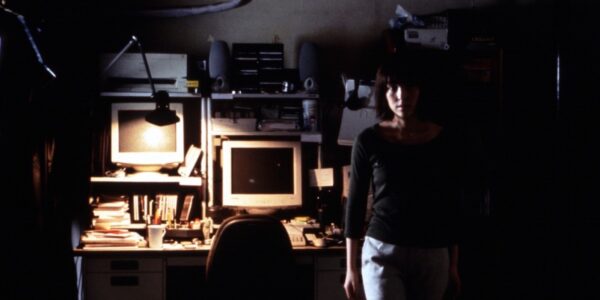 A woman leans against a computer table in a dark room looking back at camera