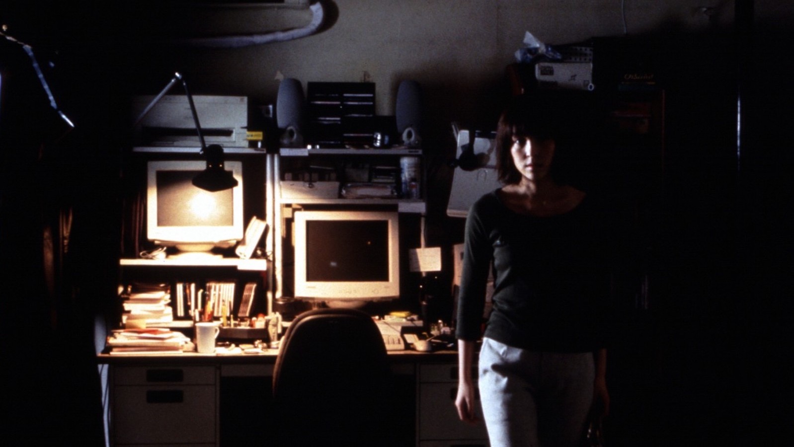 A woman leans against a computer table in a dark room looking back at camera