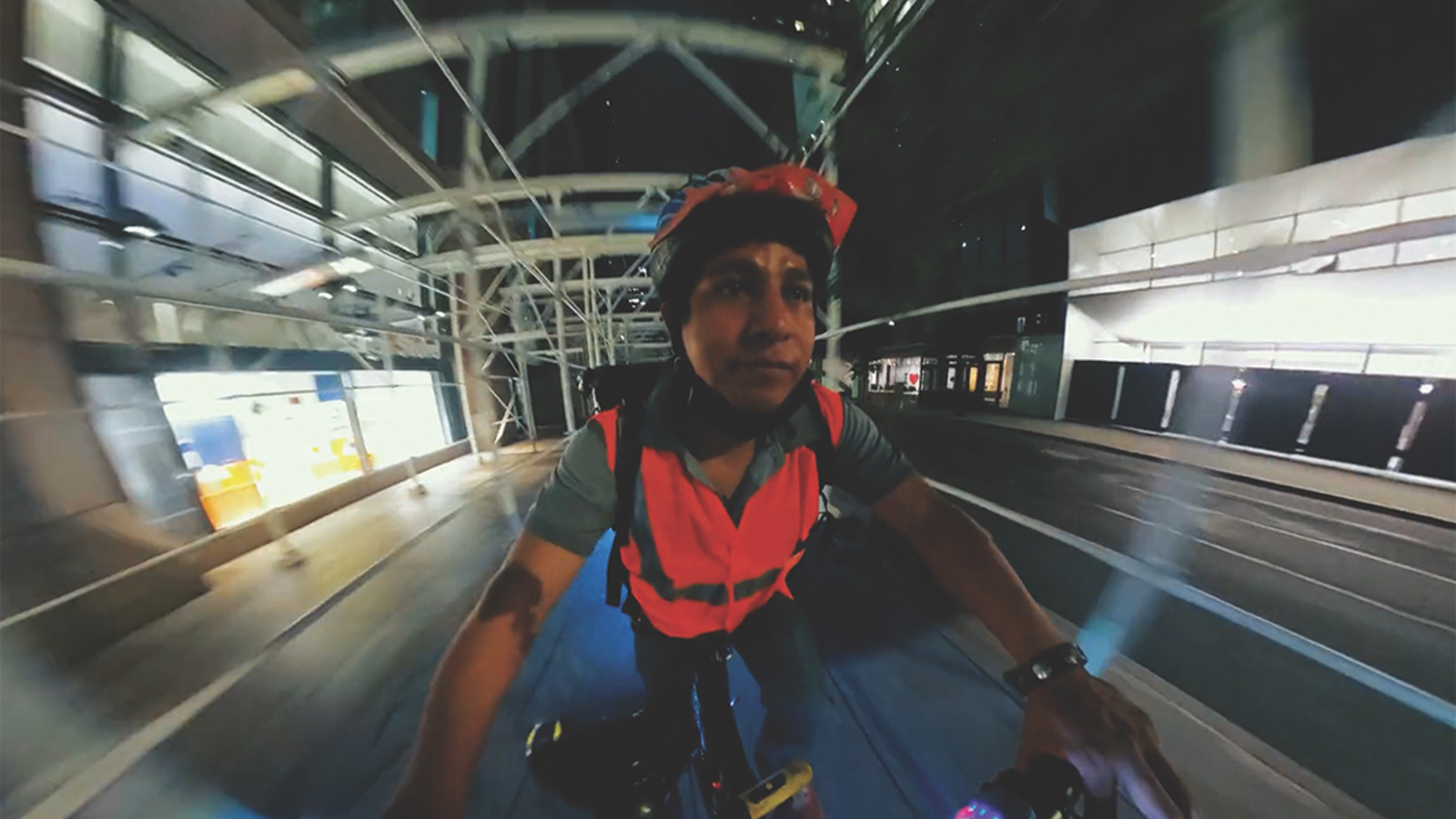 A man in a red bike helmet rides a bicycle facing camera, through city scaffolding