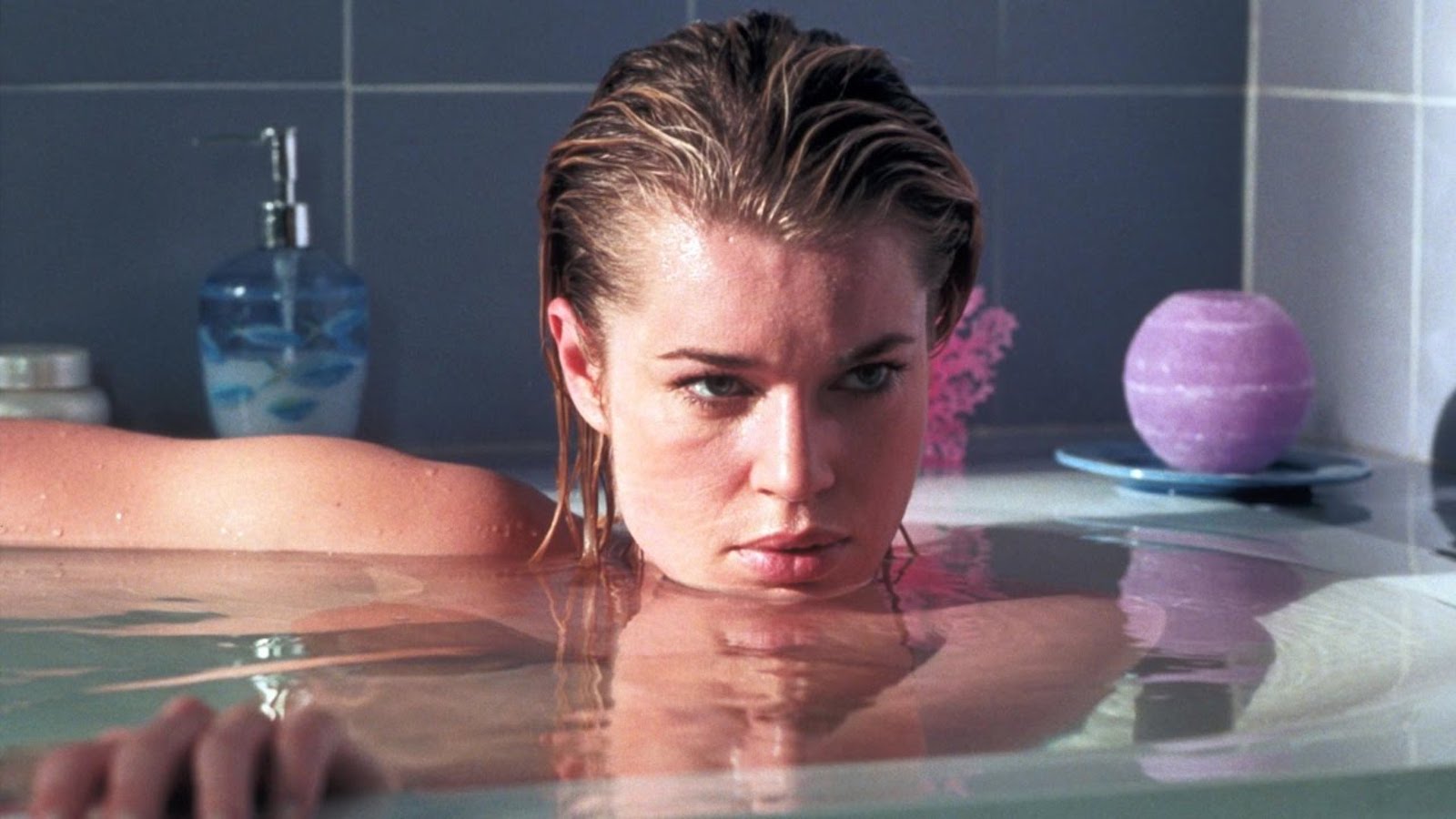 A woman sits in a bathtub, the water nearly overflowing as she looks off screen