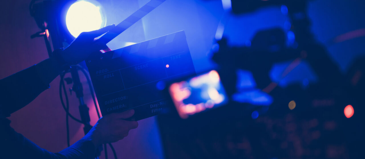 On a film stage set, a clapperboard is held up in front of a camera in front of a digital camera with light flooding from behind it.