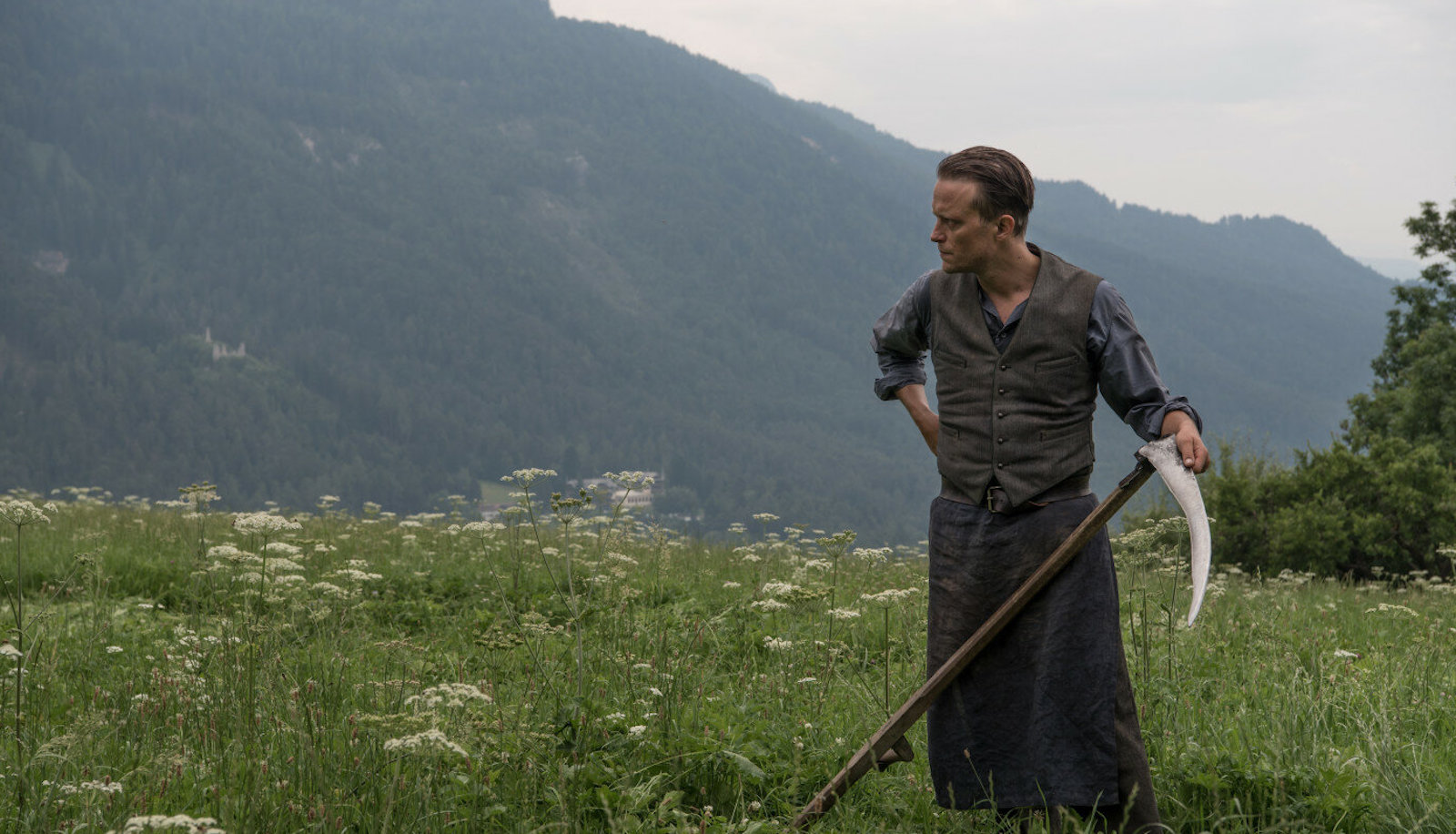 A man holds a scythe in a field of grass amidst mountains