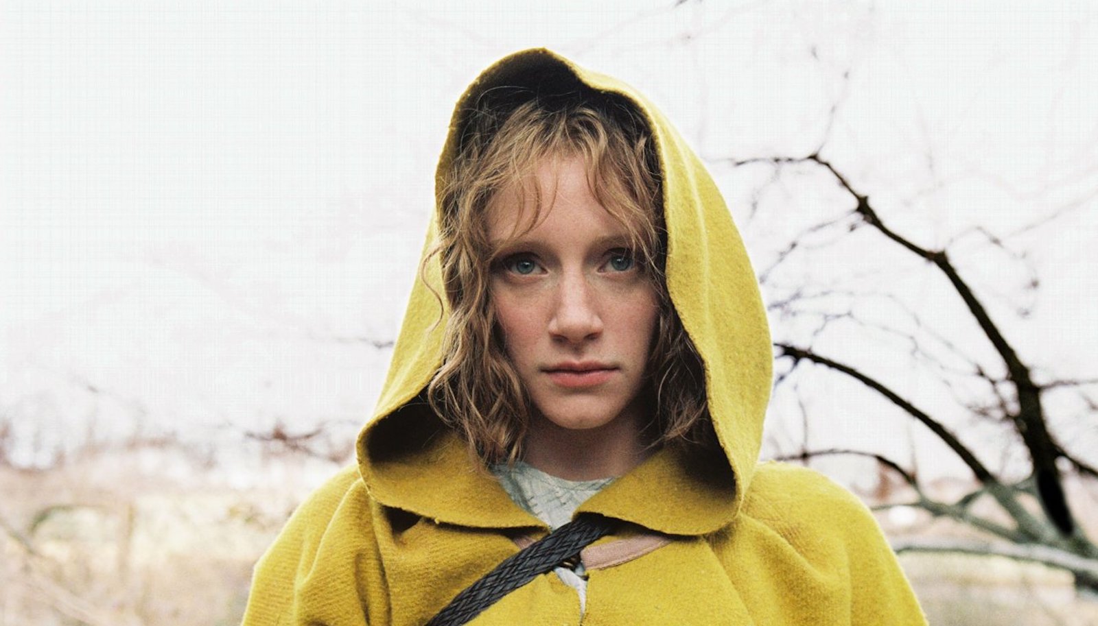 A young woman in a yellow hooded jacket looks at camera ominously in front of bare tree branches