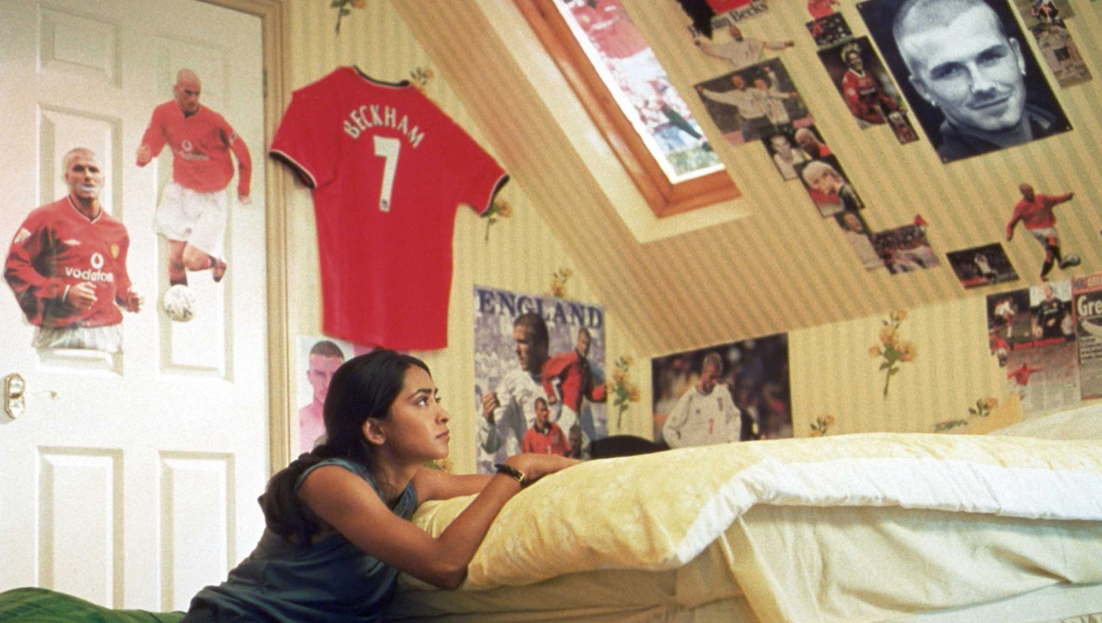 A young girl kneels by her bed and looks up at a photo of David Beckham, with images of soccer players all over her wall