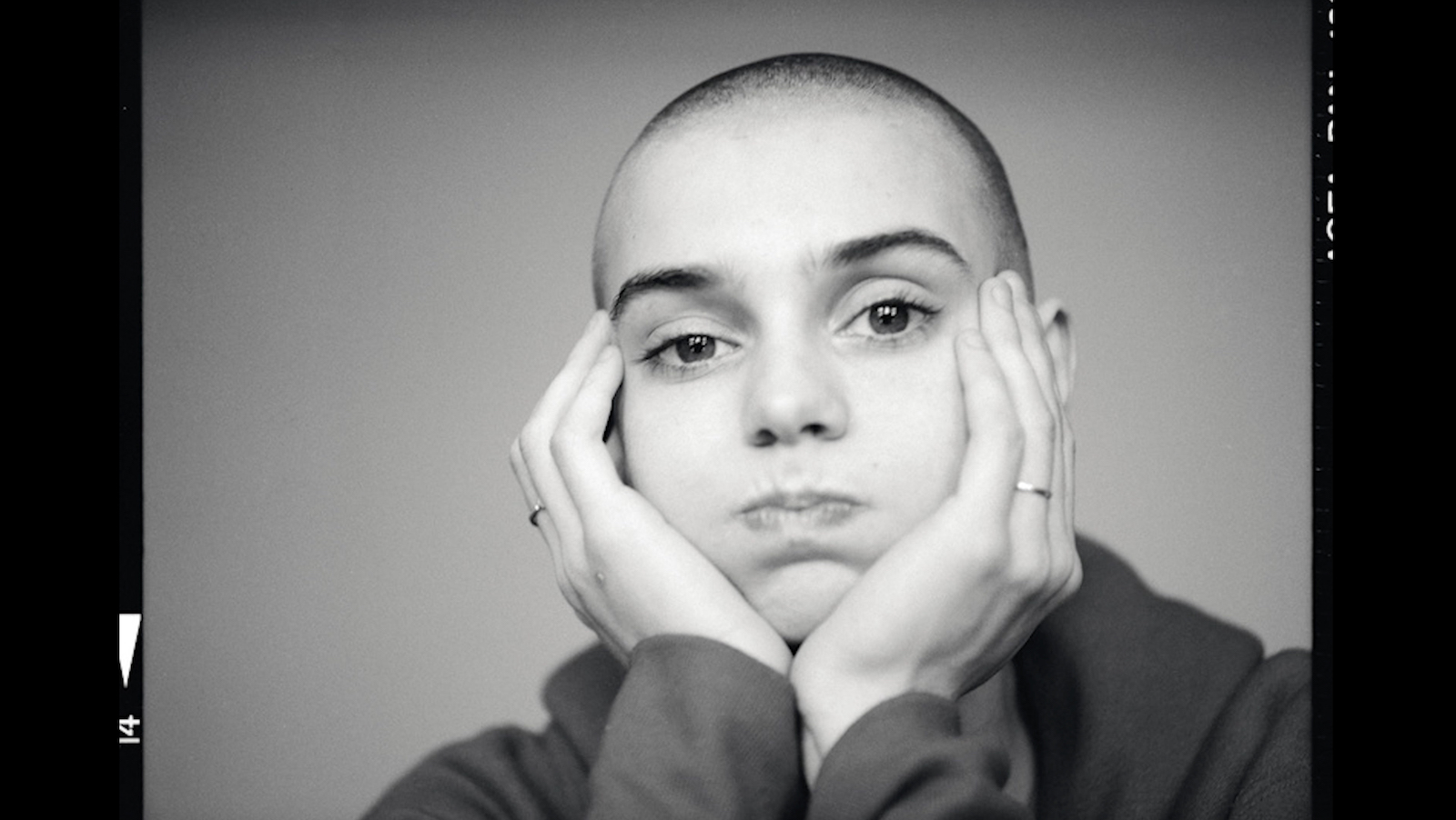 closeup on the face of a young white woman with a shaved head. Her hands frame her face.