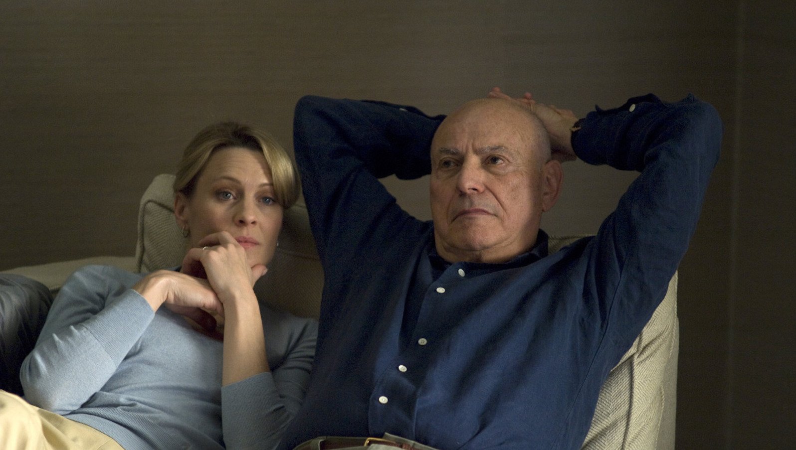 A man and woman lie in bed, both wearing blue shirts. His arms are behind his head and her hand is cradling her chin. Both are pensive.