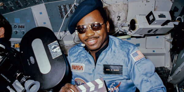 A man in hat and sunglasses and holding a film slate sits in a space shuttle smiling at camera