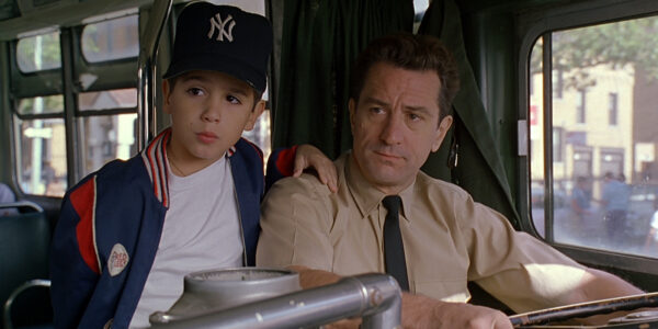 A bus driver in the driver's seat sits next to a young boy in a New York Yankees cap, who stands with his hand on the man's shoulder