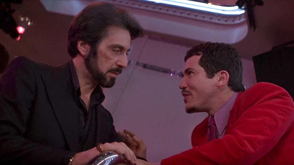 From a low angle image, a man with a beard looks intently at a shorter man in a red jacket