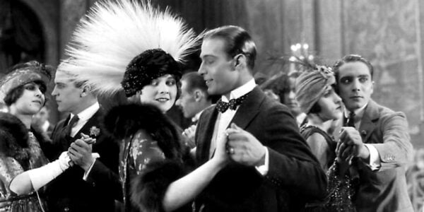 A black and white image of a man in a tuxedo dancing with a woman in a high white feathered peacock headdress