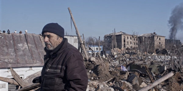 A serious looking man in a stocking cap walks before building rubble