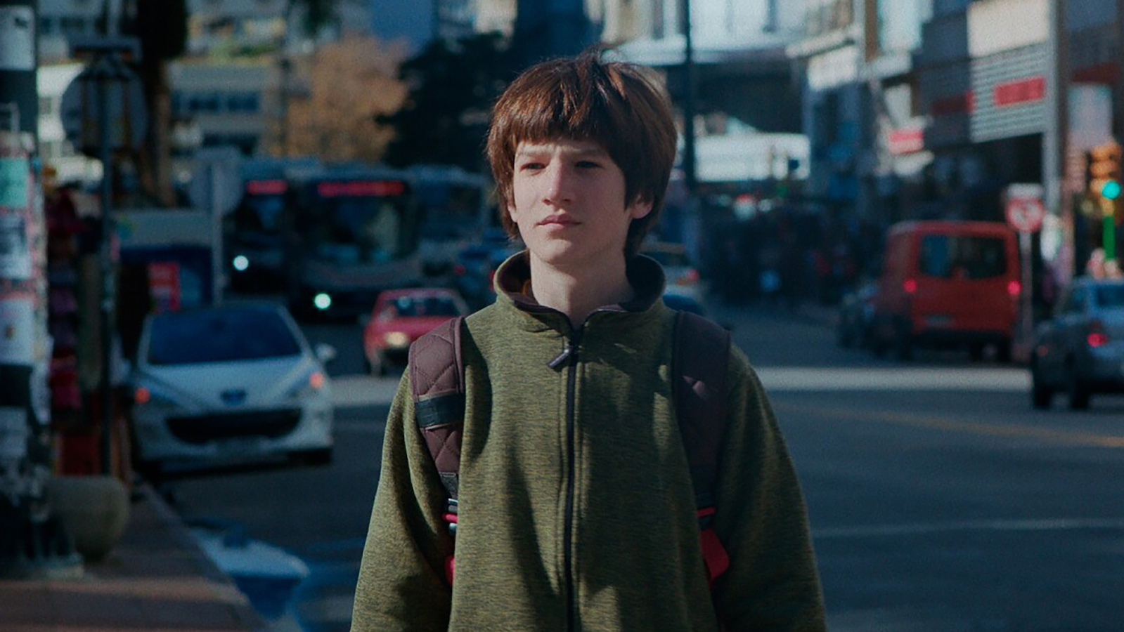 A boy wearing a sweatshirt and backpack stands in a city street and stares off