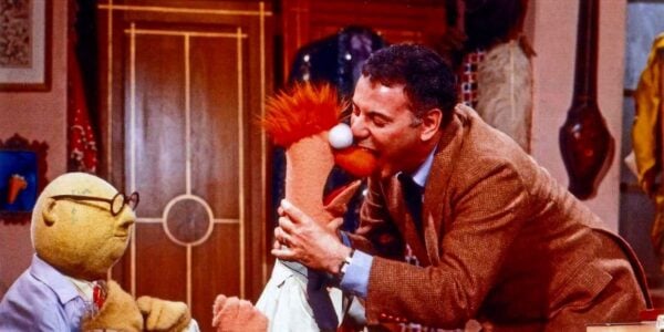 A man in a brown blazer bites a muppet on its red nose while another muppet with glasses watches.