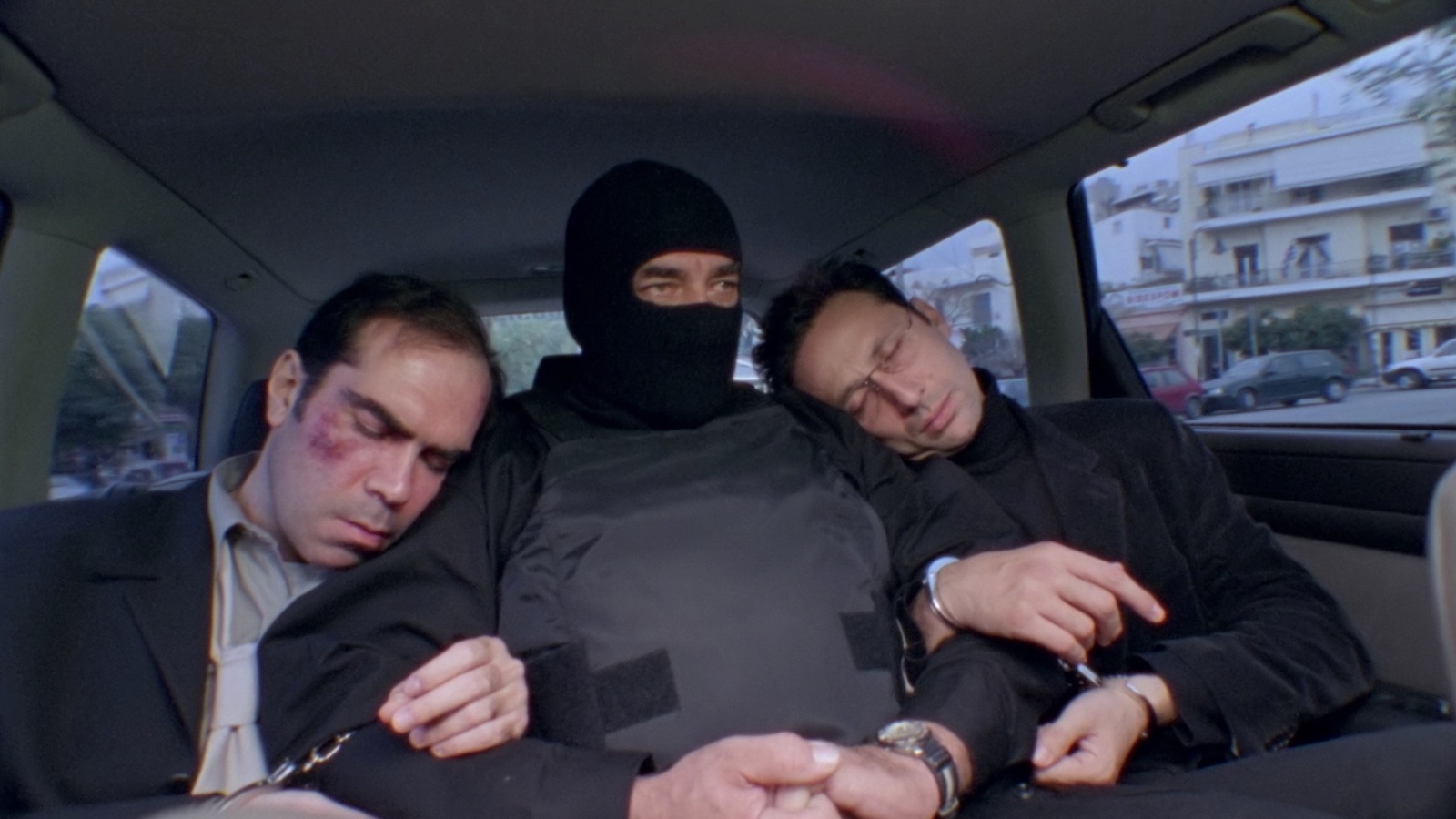 Three men sit in the back of a car, handcuffed to each other, the middle man wearing a bulletproof vest and ski mask