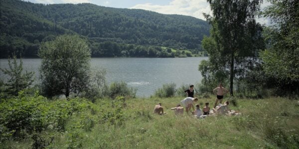 A group of people, children and adults, in bathing suits by a beautiful idyllic lake, sitting in the grass