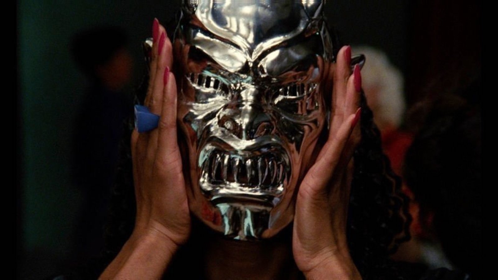 A woman with painted fingernails holds up a silver demon mask over her face and looks at camera