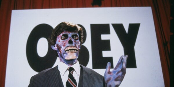 A suited man with a skeleton face stands before a large sign that says OBEY.