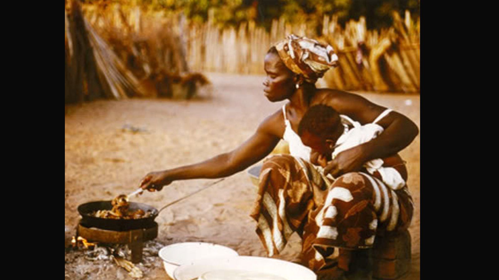 A woman holding a baby in one hand sits before an open fire cooking chicken in a rural African village