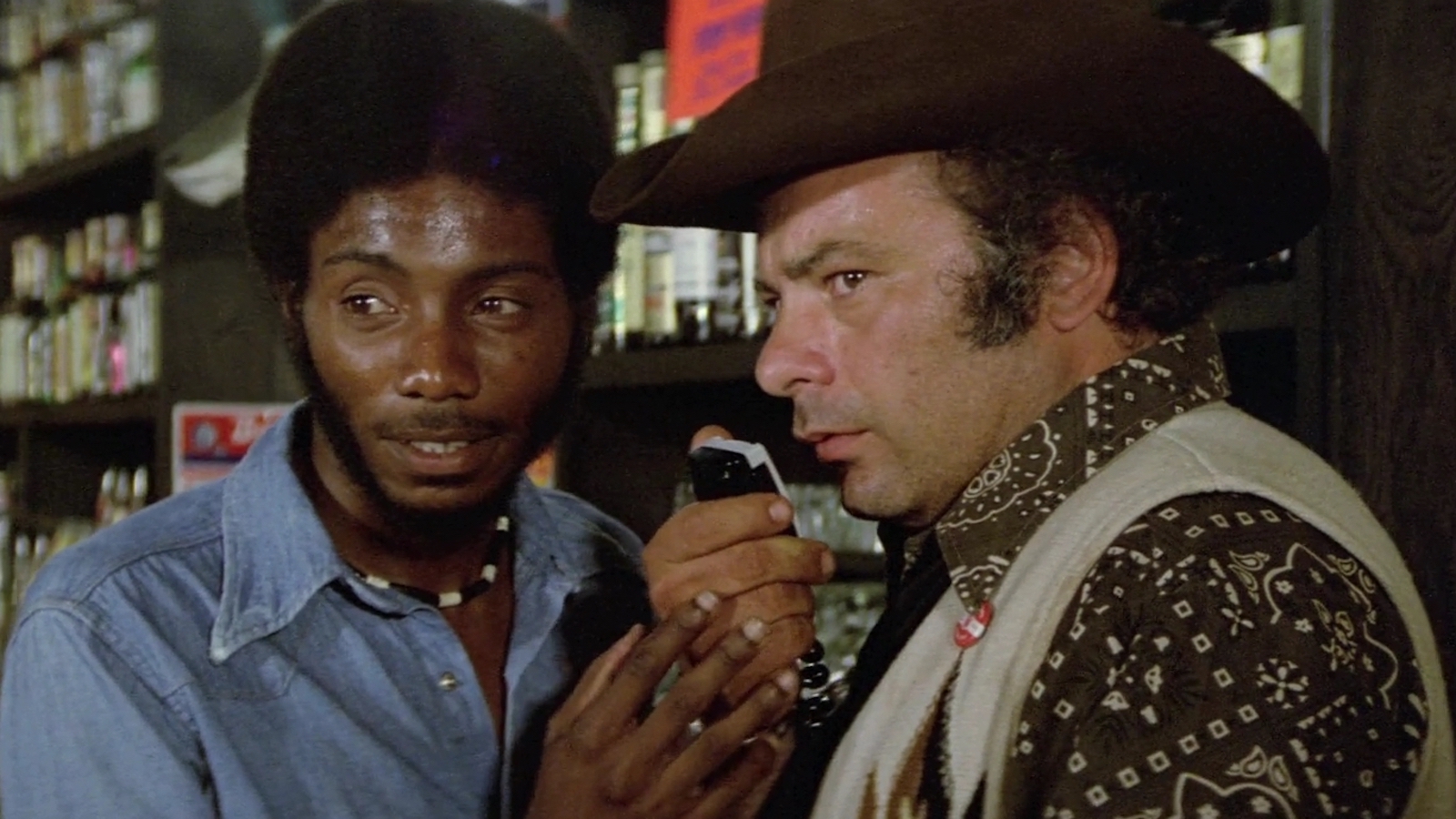 Two men stand in a liquor store, one holding a CB radio to his mouth and wearing a cowboy hat; both men look off screen with serious expressions