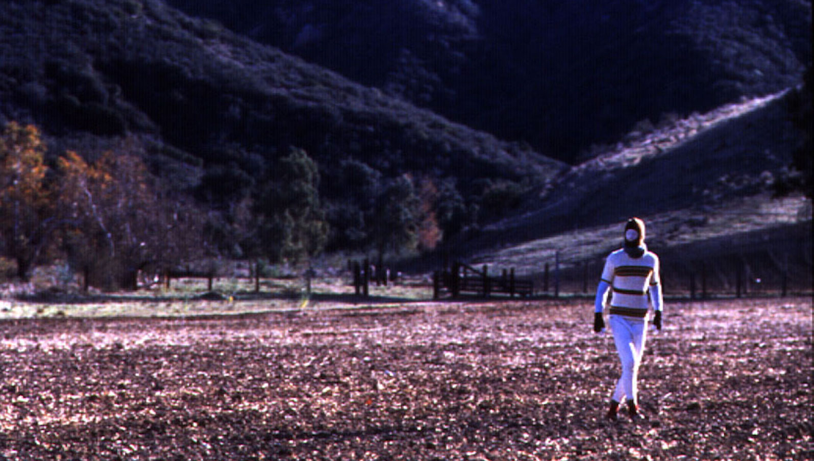 A masked figure in a white, stripe shirt and white pants walks across a vast mountainous landscape