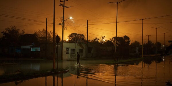A yellow sky blazes behind a flooded street at night, with a solitary figure walking through water in front of a house.