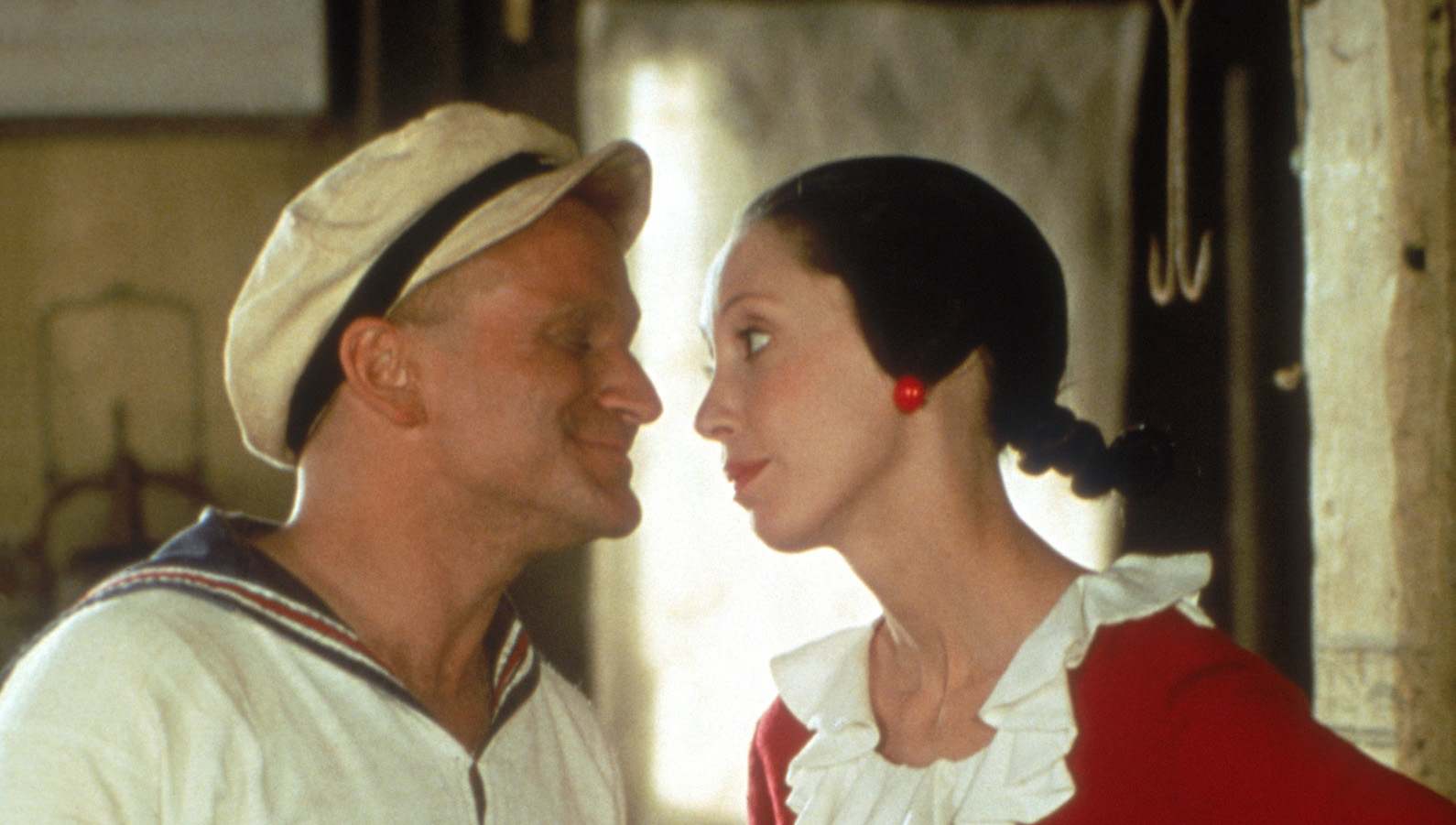 A man in a sailor suit (Popeye) and a woman with red earrings and a red dress (Olive Oyl) look each other in the eye.